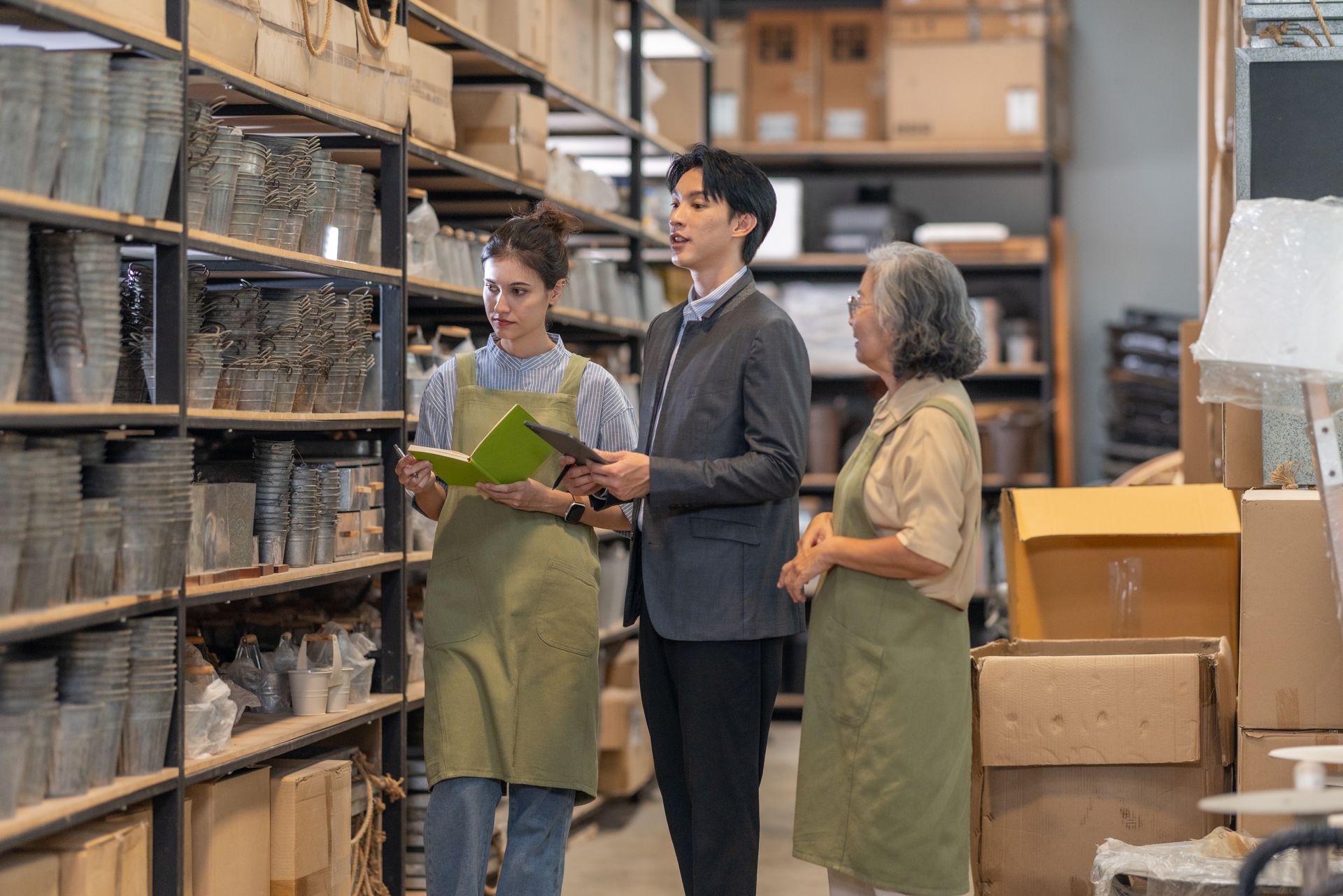 Three people in aprons reviewing a clipboard in a warehouse aisle lined with shelves and boxes