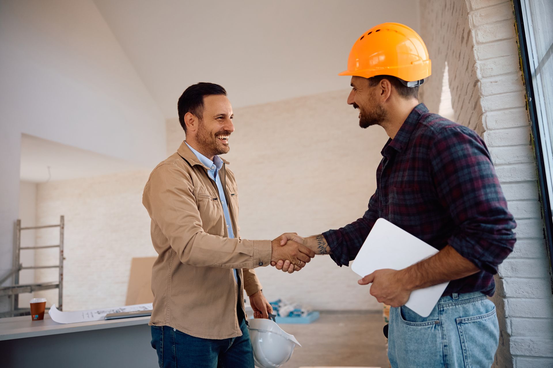 Two men shaking hands at a construction site, one wearing an orange hard hat and holding papers.