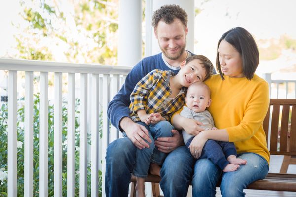 Family of four sitting on a porch bench, smiling and holding a baby in bright daylight