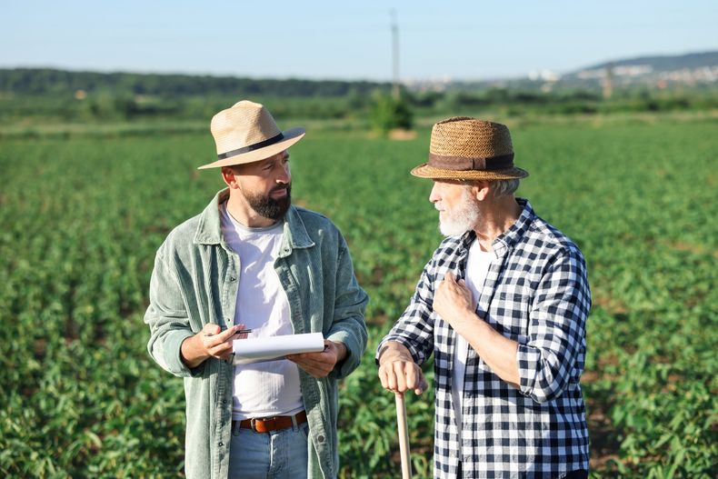 Two farmers in straw hats talk in a green field, one holding a clipboard and the other a walking stick.