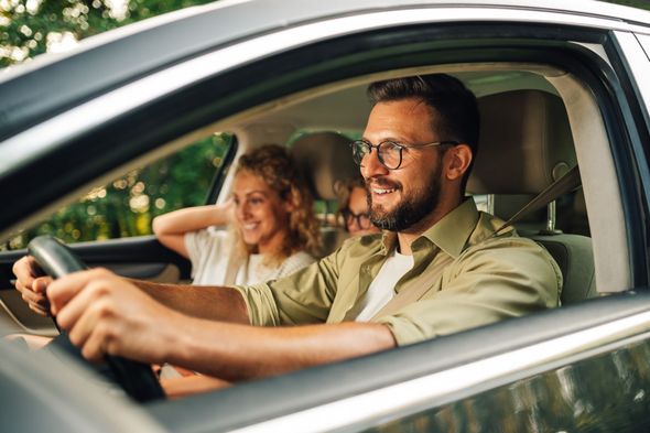 Man driving a car with a smiling passenger in the back seat, seen through the open window.