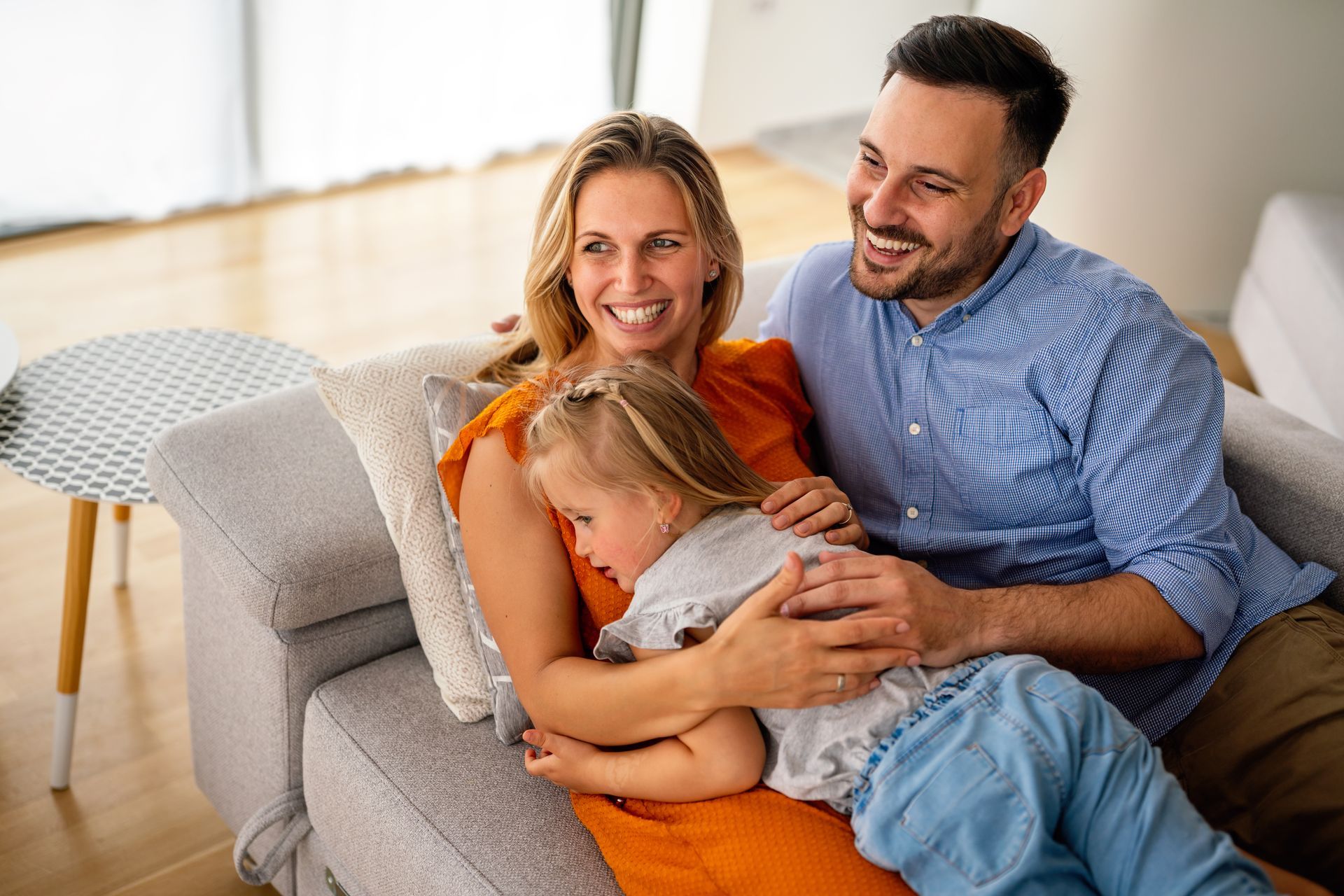 Family cuddling on a couch, smiling in a bright living room, with a child resting on an adult’s lap
