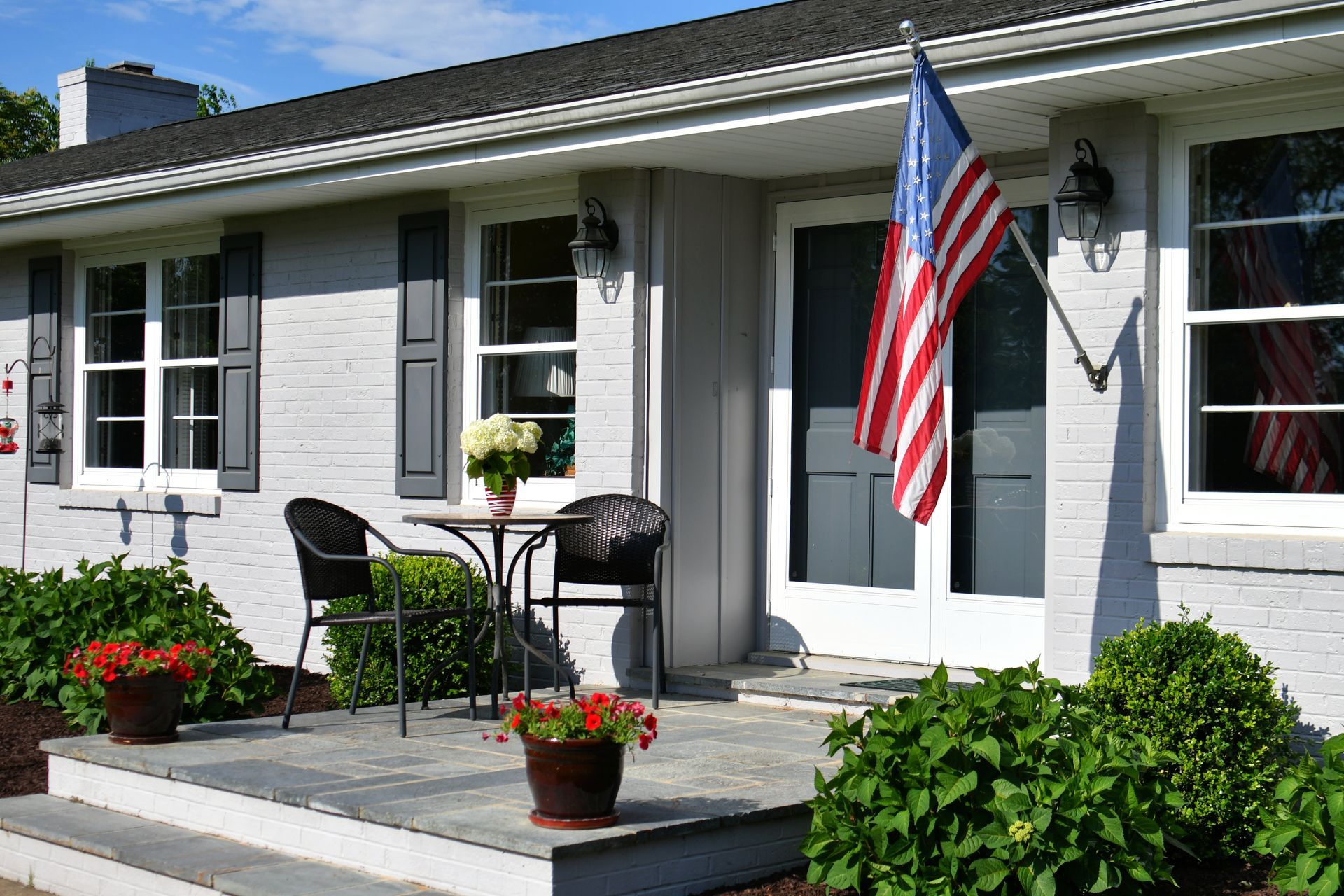 Suburban house front porch with American flag, chairs, and potted flowers in sunlight