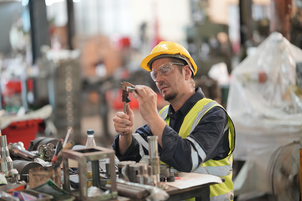 Worker in a yellow hard hat and safety vest using pliers at a factory workbench