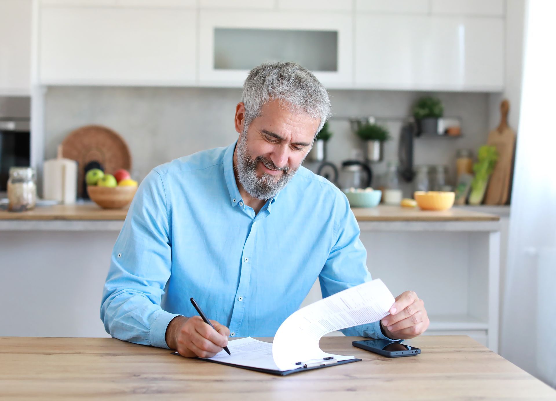 Man in a blue shirt signing papers at a kitchen table with a phone beside him