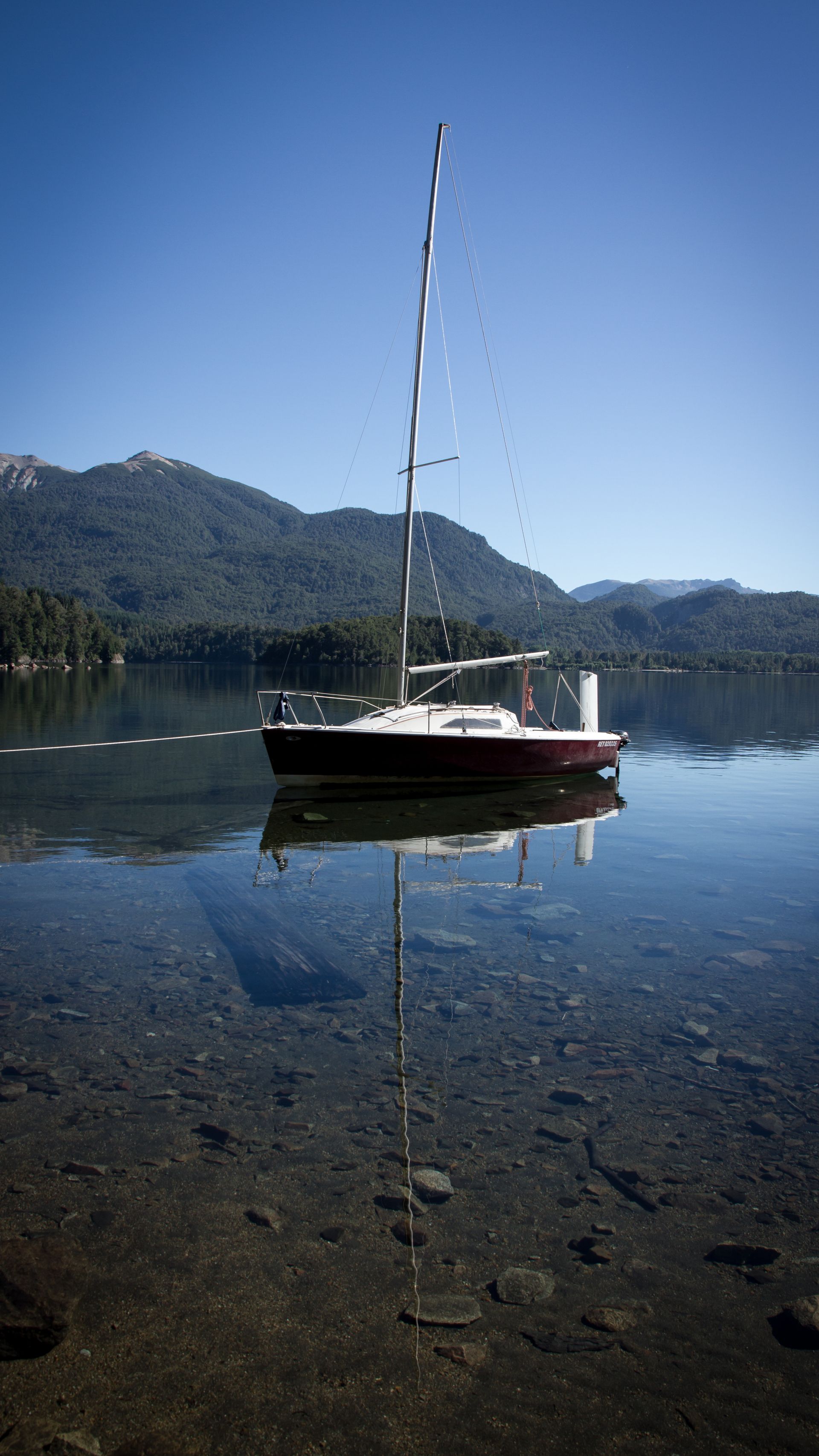 Small sailboat on a calm lake with mountains reflected in the water under a clear blue sky