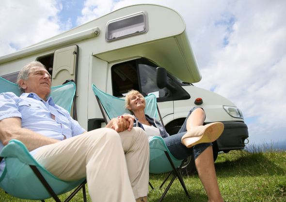 Two people relaxing in folding chairs beside a green-and-white camper in a grassy field