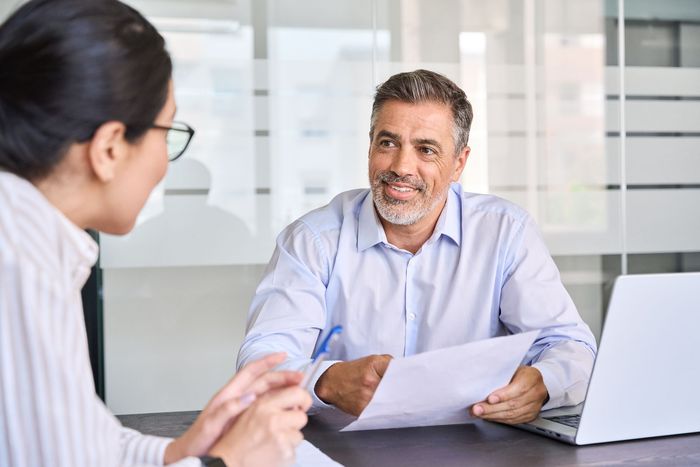 Two coworkers in a bright office discussing documents across a table, with a laptop nearby.