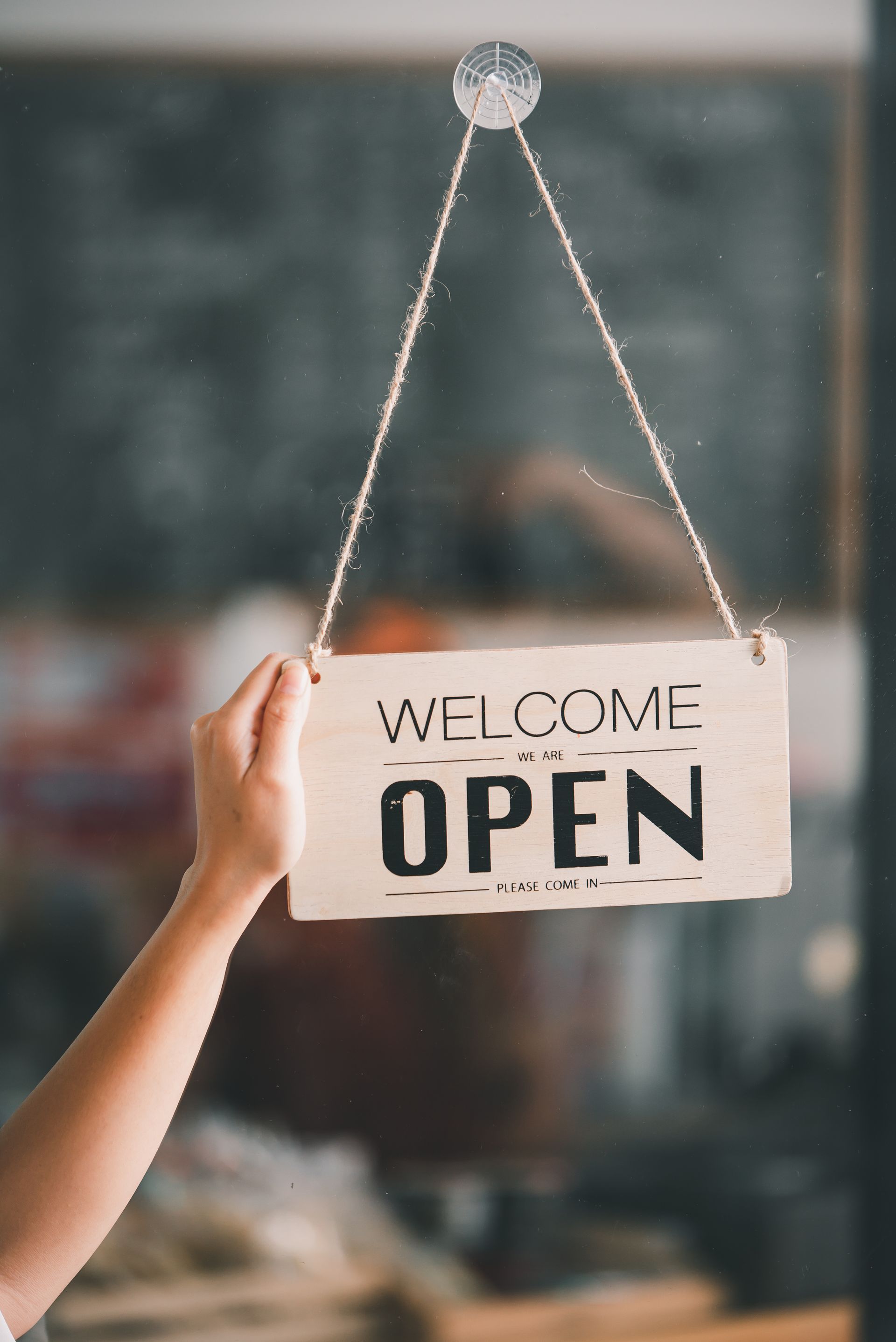 Hand holding a hanging sign that says “WELCOME OPEN” in a blurred storefront window.