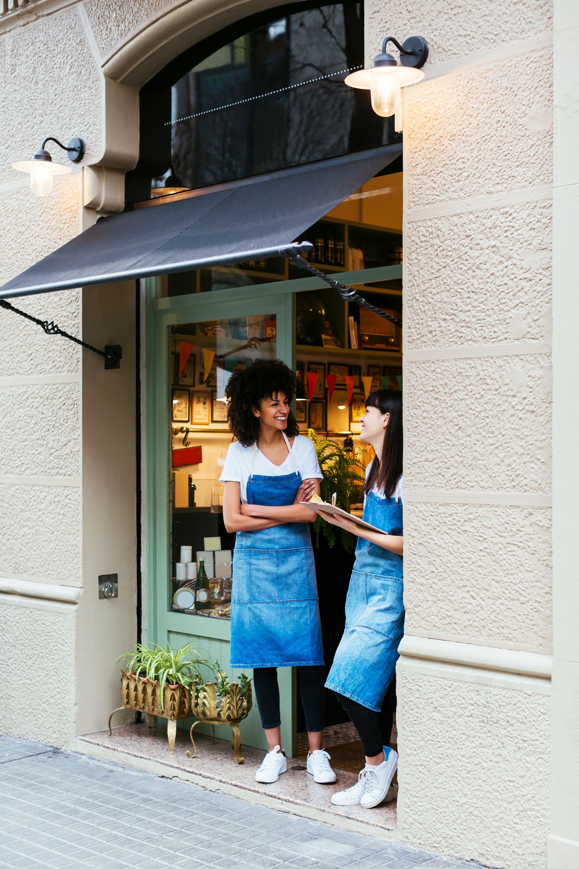 Two baristas in blue aprons stand in a café doorway holding drinks