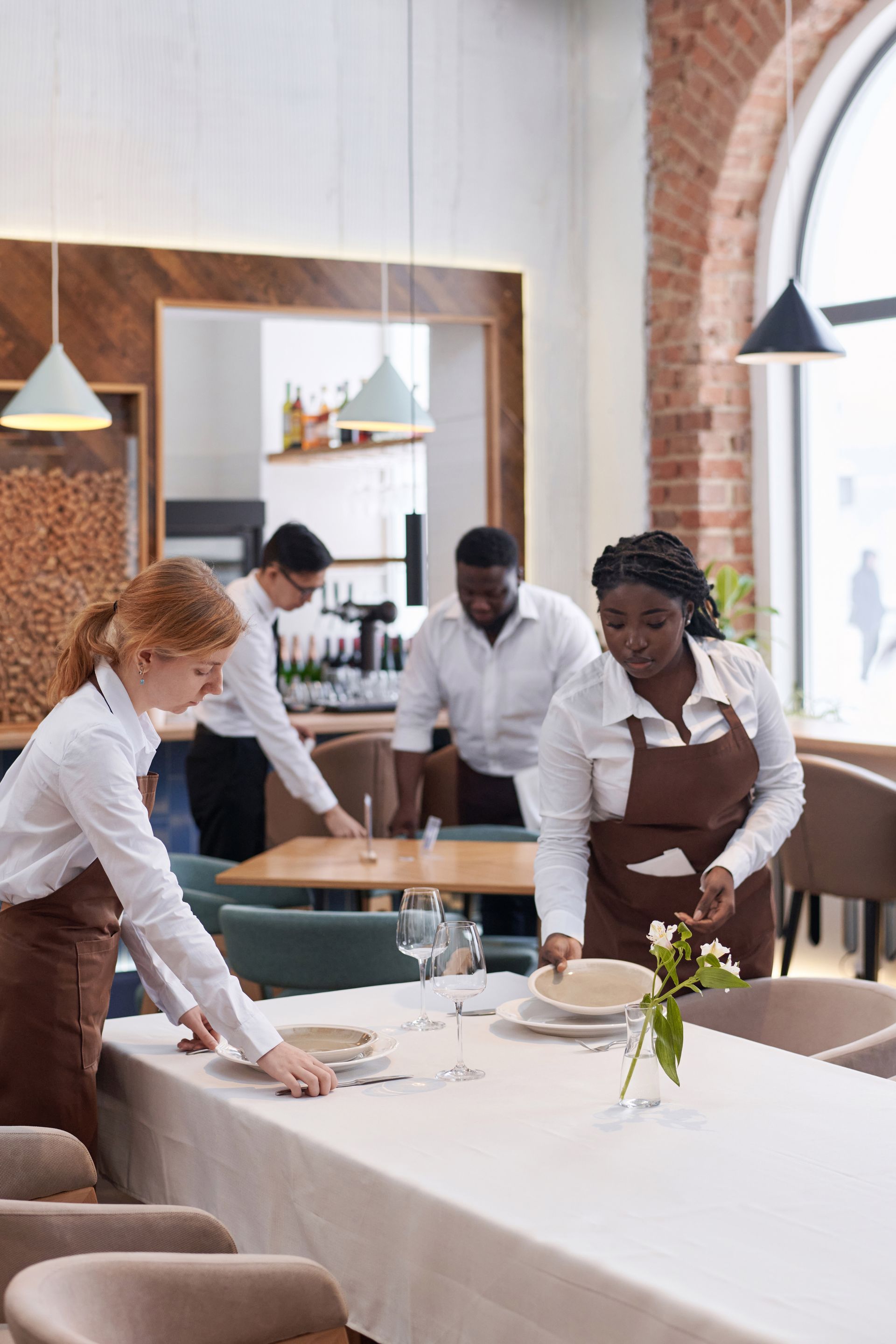 Staff setting a dining table in a bright restaurant with white tablecloths and hanging pendant lights