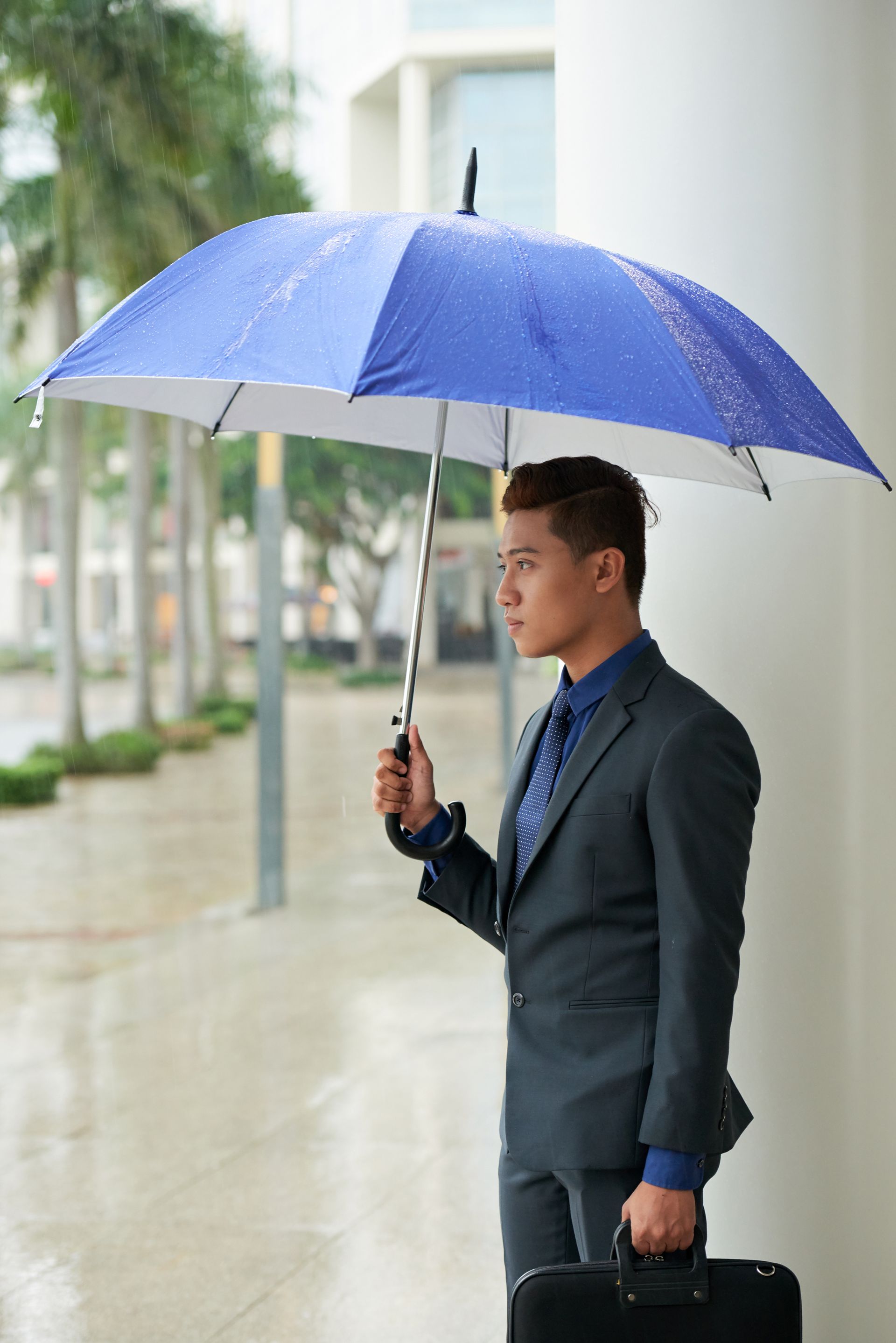 Man in a dark suit holding a blue umbrella while standing outdoors in the rain
