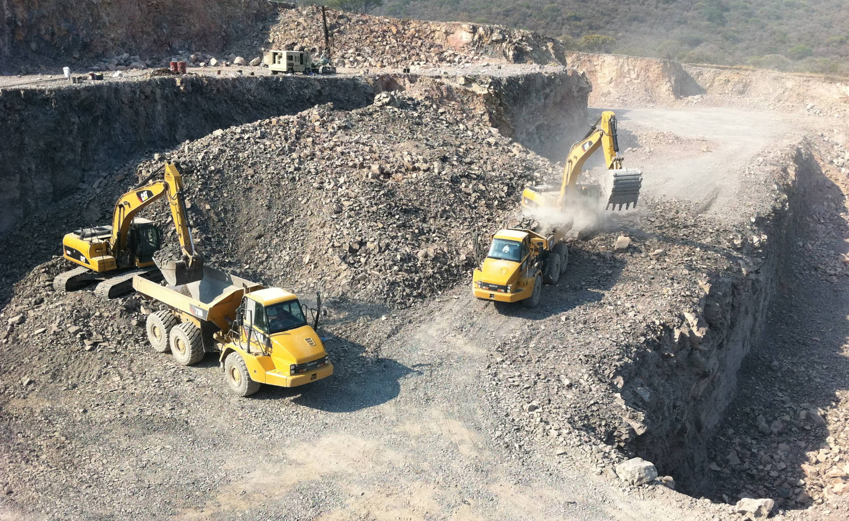 Vehículos de construcción amarillos en una cantera: excavadoras cargando un camión volquete. Entorno polvoriento y rocoso.