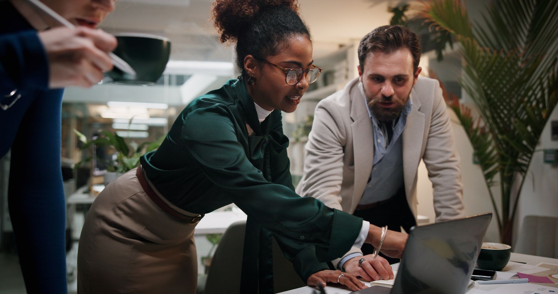 Two coworkers lean over a laptop in a bright office, pointing and discussing something on screen.