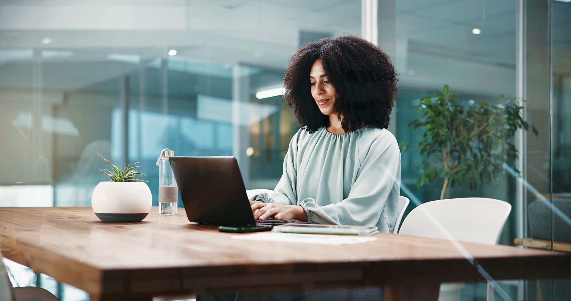 Woman working on a laptop at a wooden table in a bright modern office with plants.