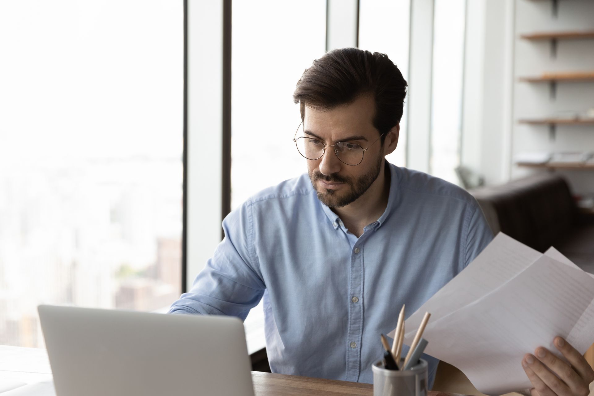 Man in glasses working at a laptop and reviewing papers at a desk in a bright office