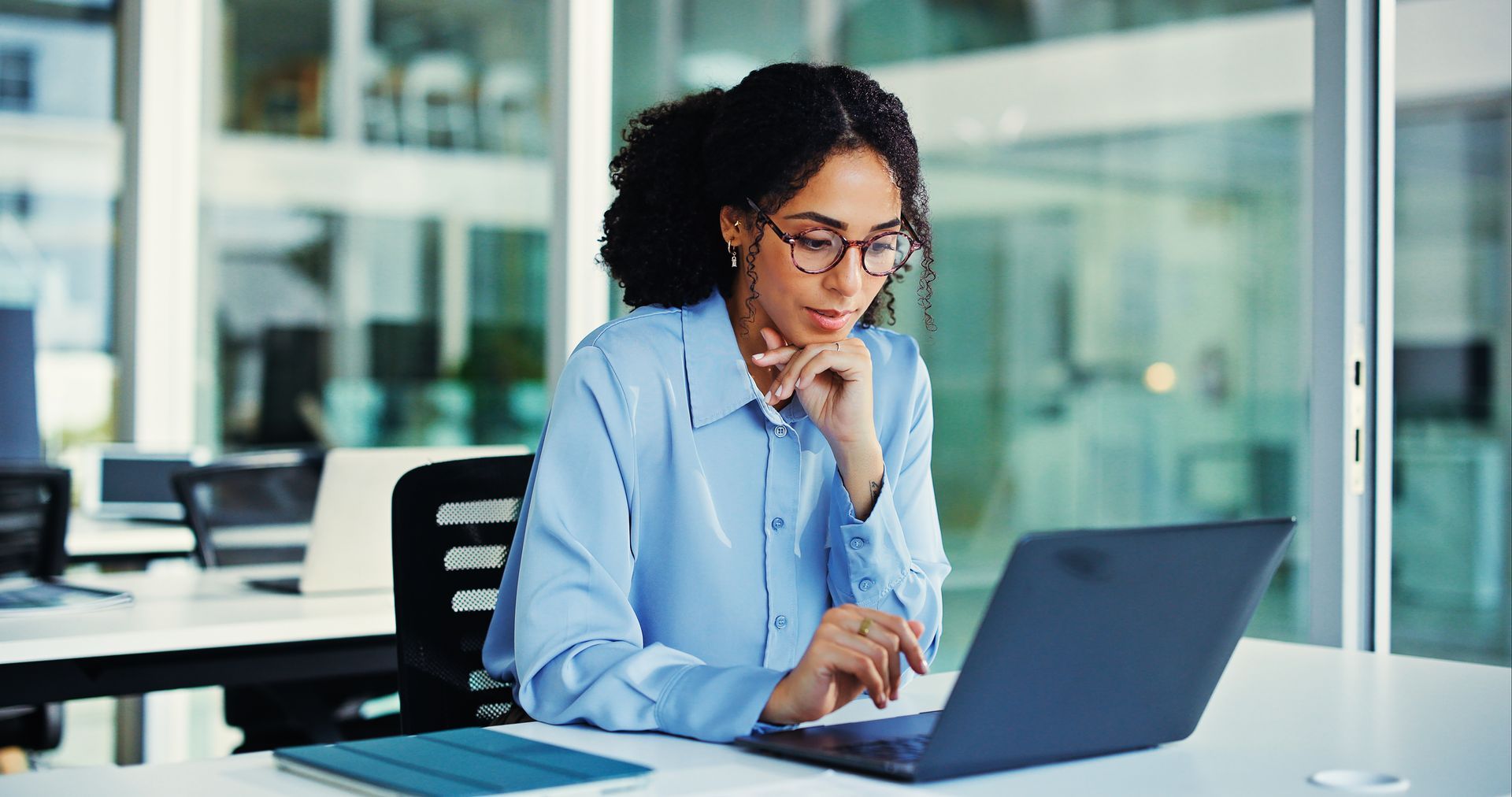Woman in a light blue shirt working on a laptop in a bright office, focused and taking notes.