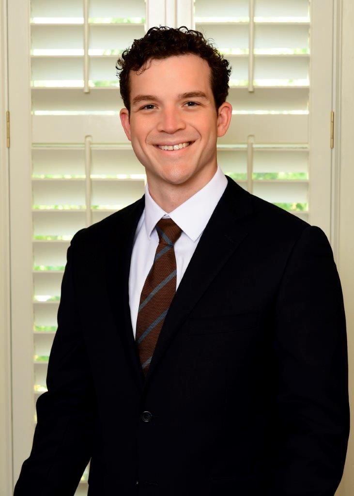 Mac Casey in a dark suit and tie standing in front of white window shutters