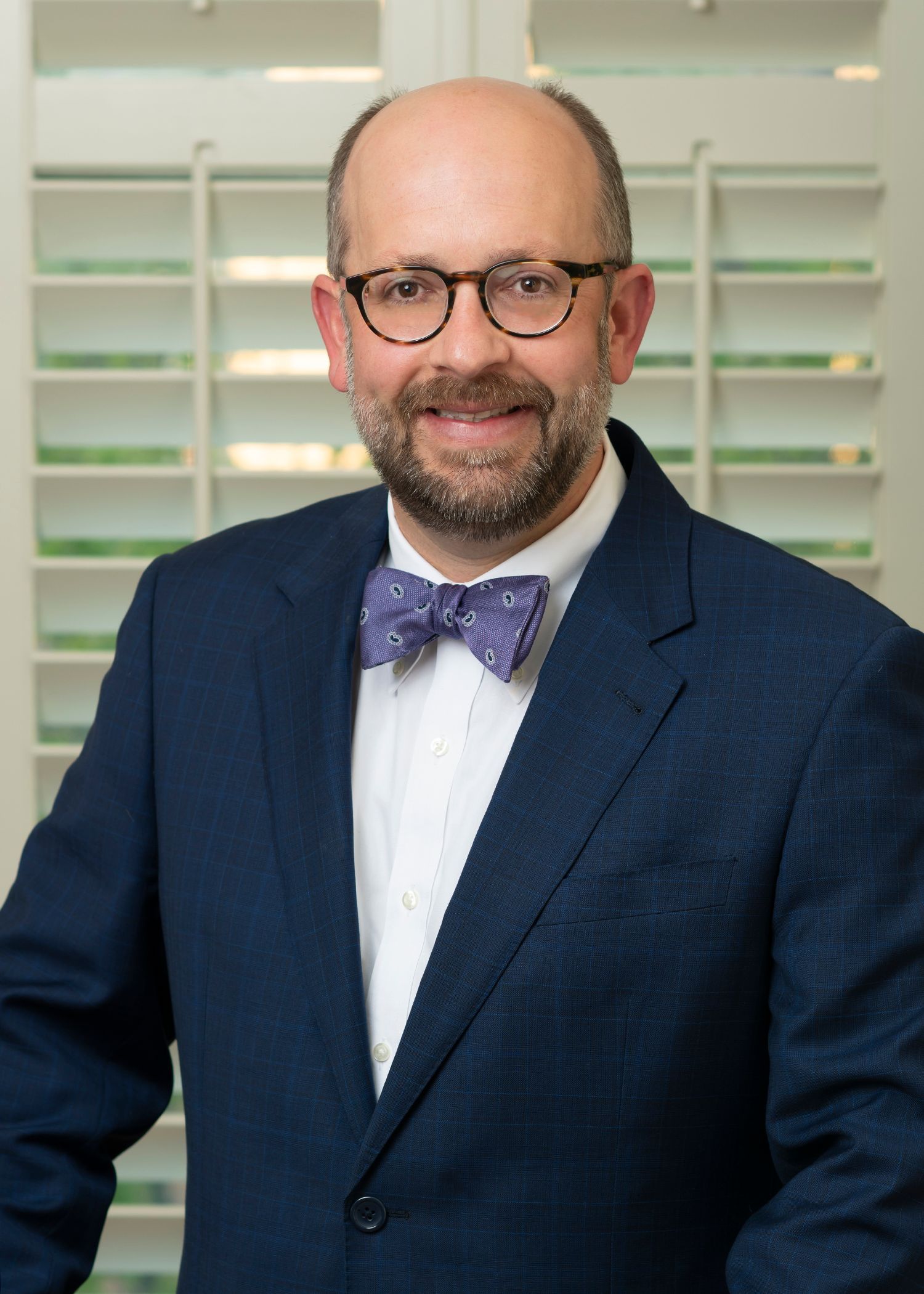 A man in a suit and bow tie is standing in front of shutters.