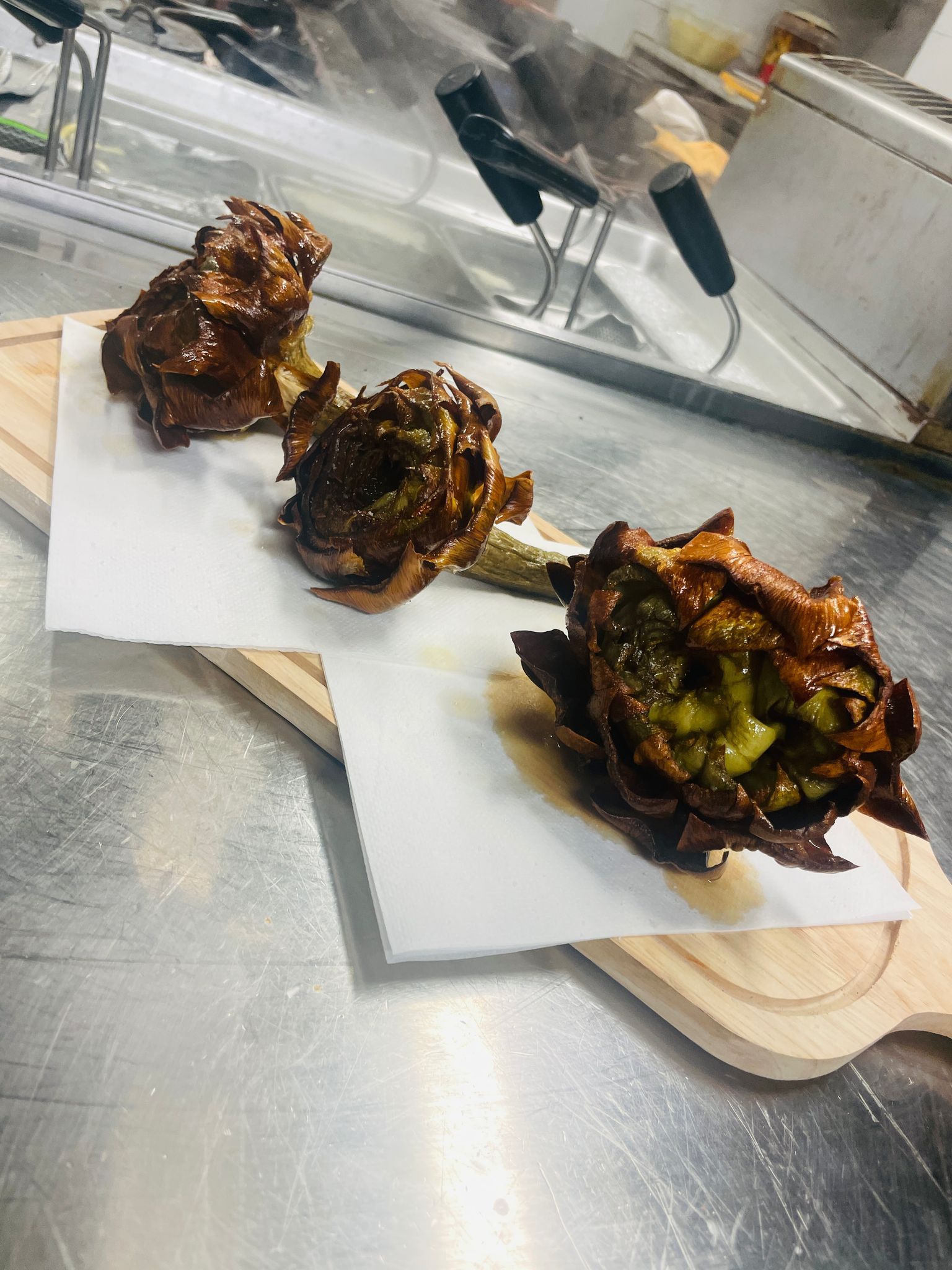 Tre carciofi fritti su taglieri di legno, presentati su carta bianca, su una superficie di acciaio inossidabile.