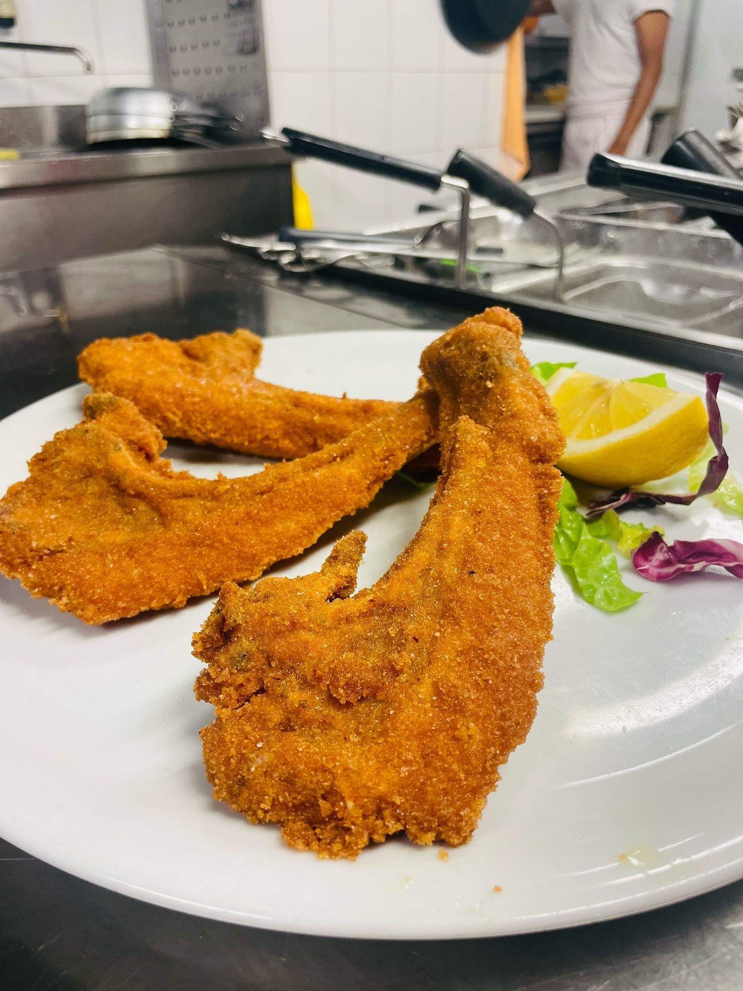 Costolette di vitello fritte e impanate su un piatto bianco con contorno di limone e lattuga. Ambiente cucina.