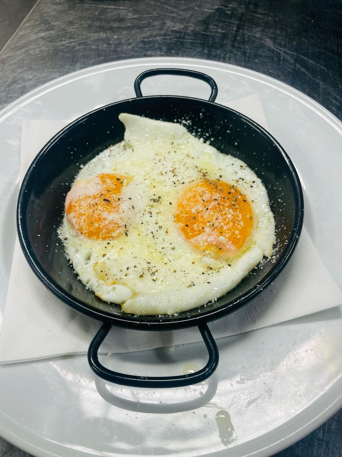 Due uova fritte in una padella nera, cosparse di formaggio e pepe, su un piatto bianco.