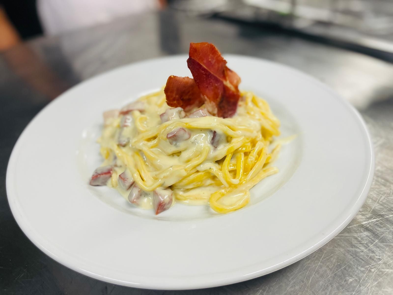 Spaghetti alla carbonara con guarnizione di pancetta su un piatto bianco, appoggiato su una superficie in acciaio inox.