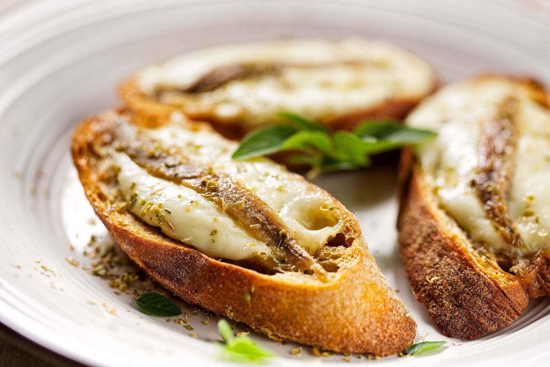 Fette di pane tostato condite con formaggio fuso e acciughe, guarnite con erbe aromatiche.
