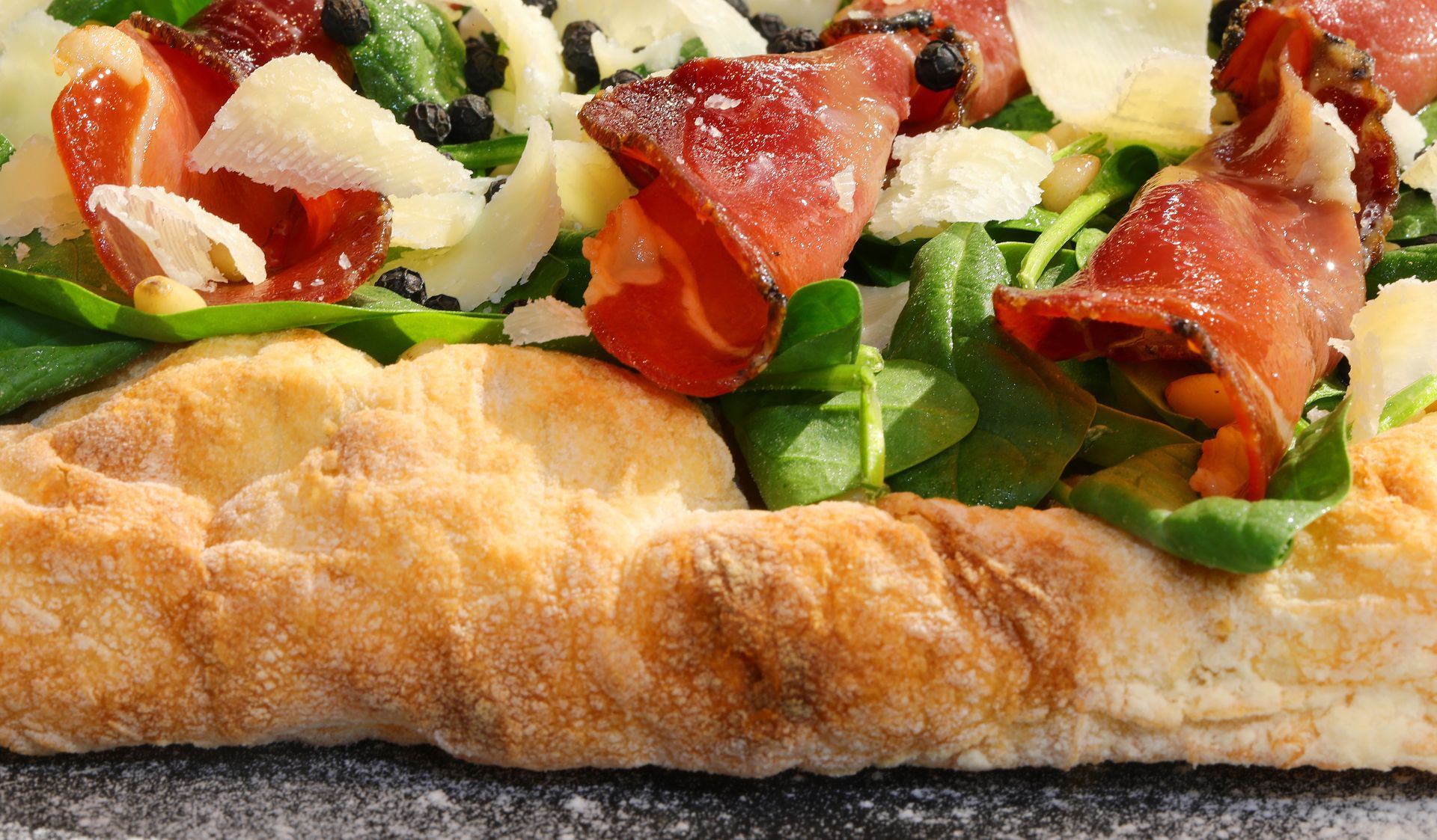 Primo piano di una pizza piatta condita con prosciutto, rucola, parmigiano grattugiato e pepe.