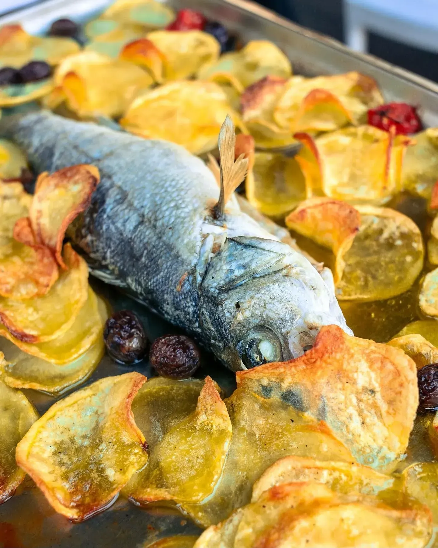 Pesce intero al forno circondato da patatine dorate, olive e pomodori.