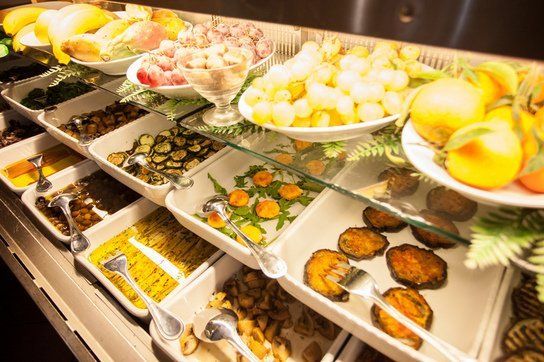 Buffet con vari piatti colorati, tra cui verdure, frutta e cibi pronti, esposti in una refrigerata vetrina di vetro.