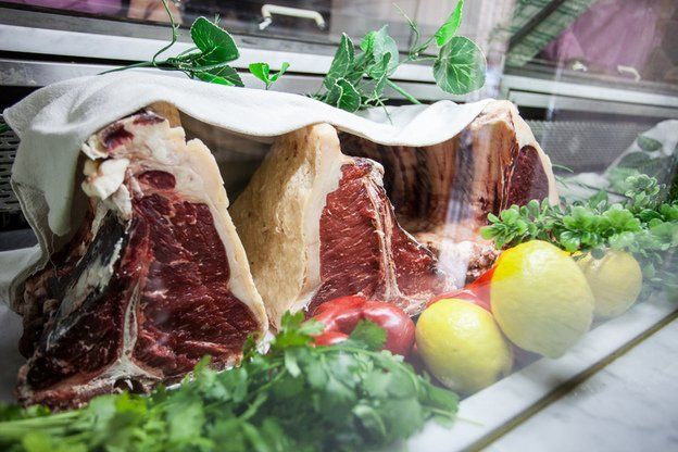 Esposizione di grandi bistecche T-bone crude sotto un telo bianco, con limoni, pomodori e prezzemolo.