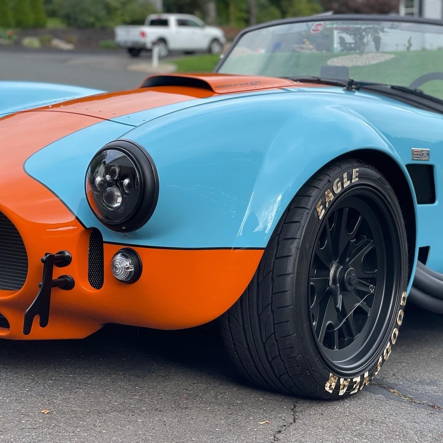 Blue and orange Shelby Cobra roadster with black wheels, parked on asphalt.