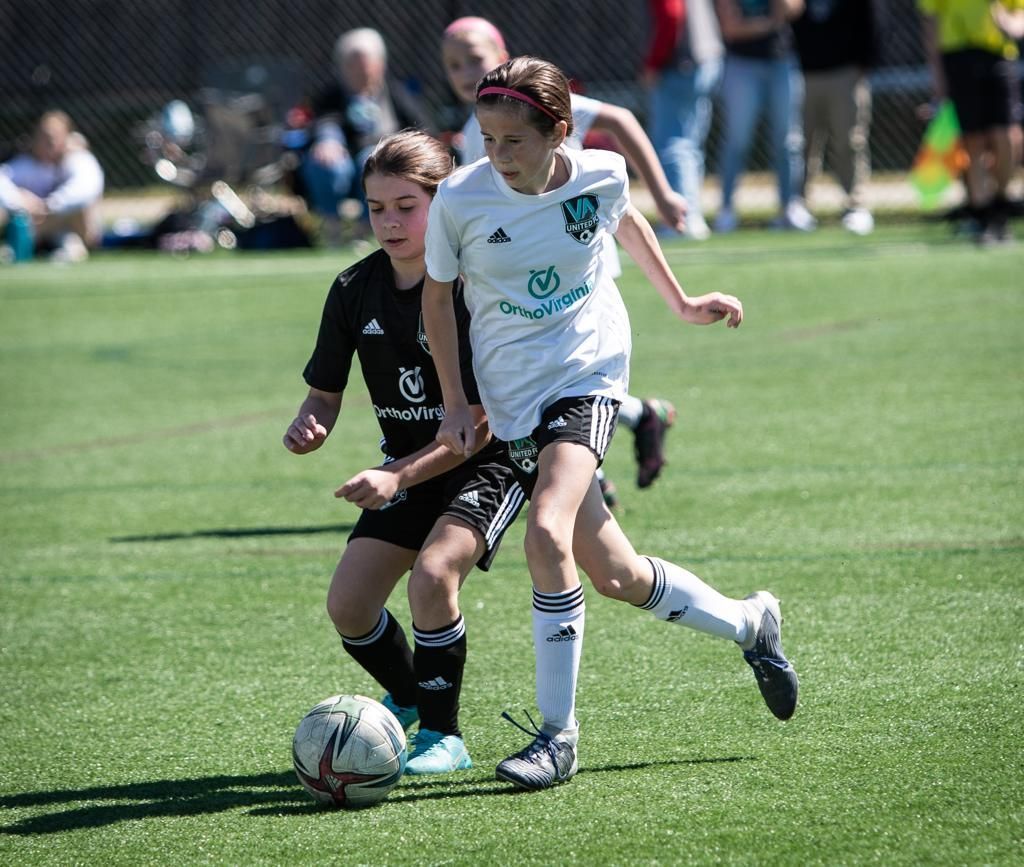 Two soccer players in action on a green field. One in black, the other in white, both focused on the ball.