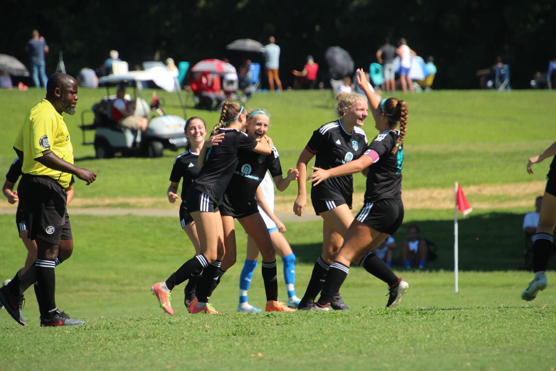 Soccer players in black uniforms celebrating on a green field; referee watches.
