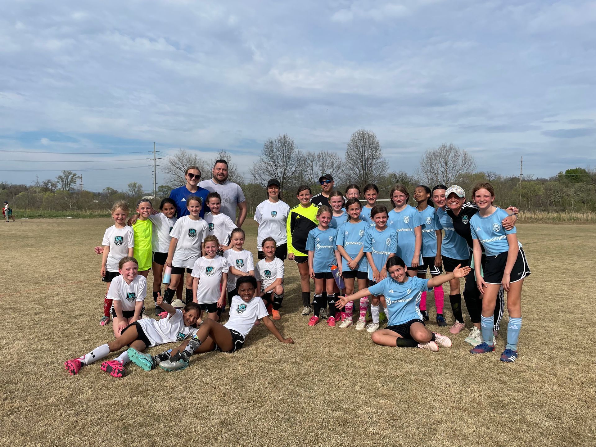 Soccer team poses on a field; wearing light blue and white uniforms. Two adults stand behind the children.