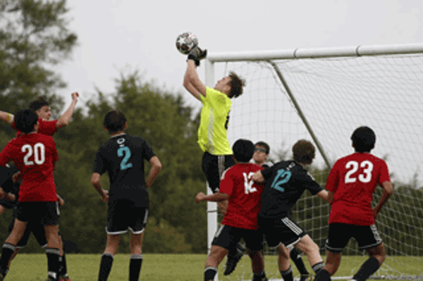 Soccer goalie in neon yellow uniform catches the ball in mid-air, surrounded by players in red and black jerseys.