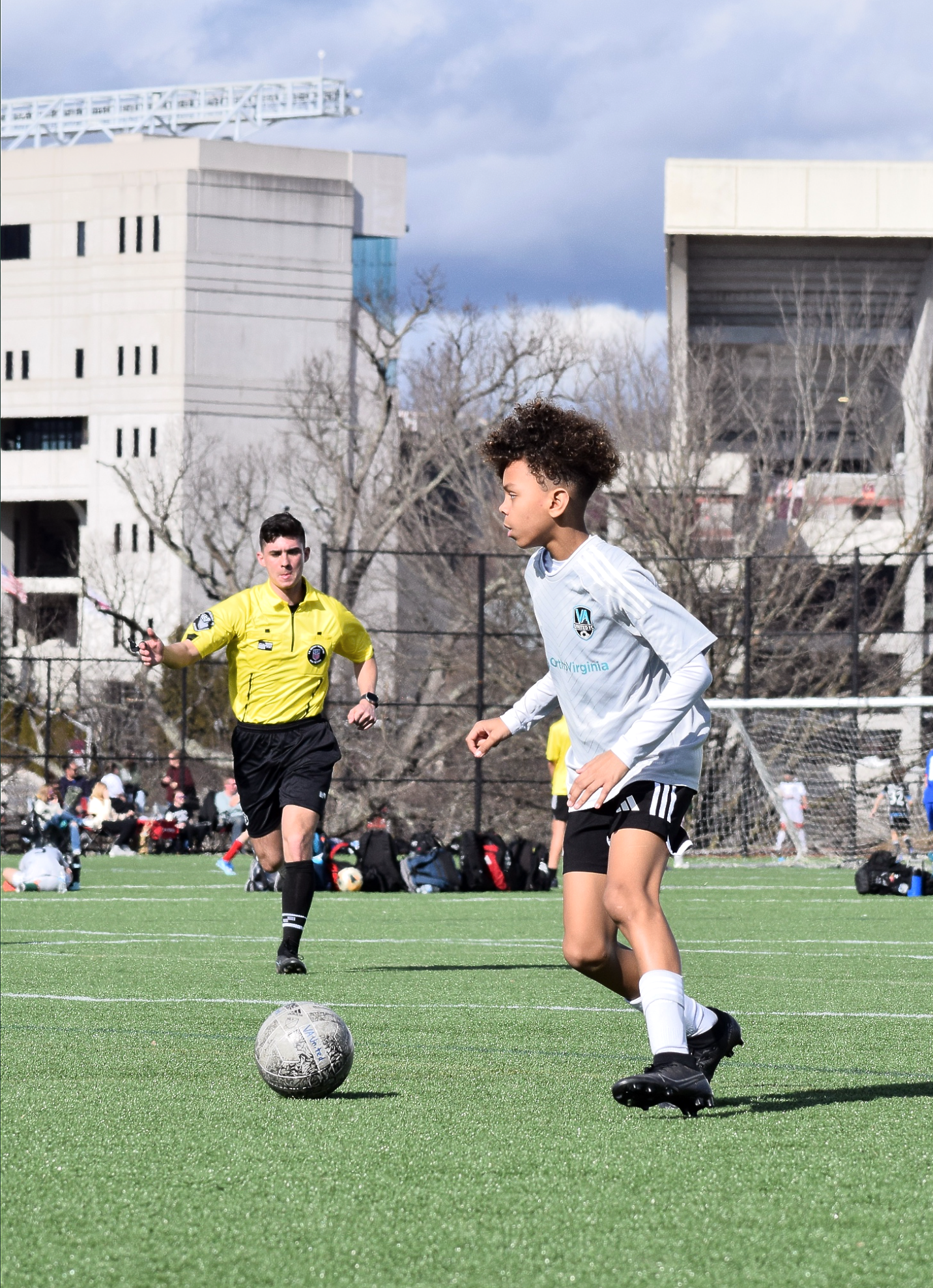 Boy dribbles soccer ball on green field as referee runs with arm raised; outdoor setting.
