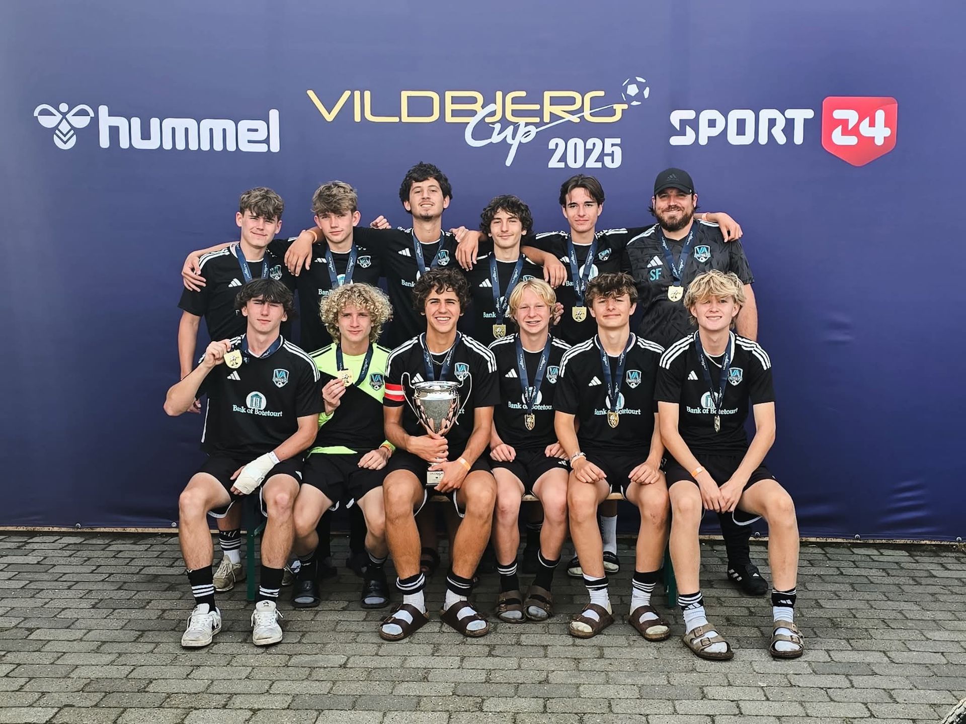 Soccer team holding trophy, wearing black uniforms, posing on blue background with logos.
