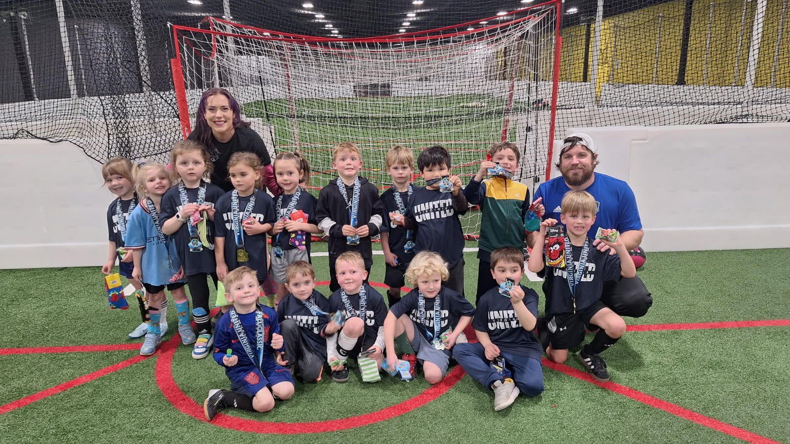 Youth soccer team posing with coaches in front of a goal on a green indoor field. Many wearing medals.