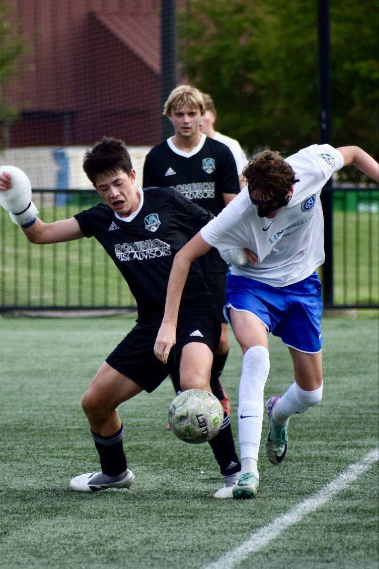 Soccer players in black and white uniforms compete on a green field. One kicks the ball, the other tries to block.
