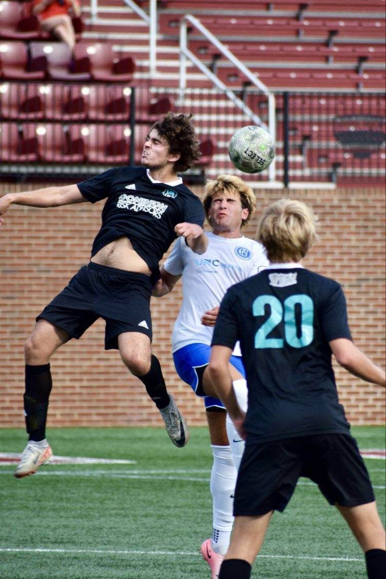 Soccer players in action, one leaping to head the ball. Game played on a green field, stadium in the background.