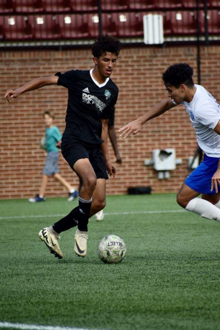 Soccer player in black uniform dribbles ball, opposing player reaches. On a green field.