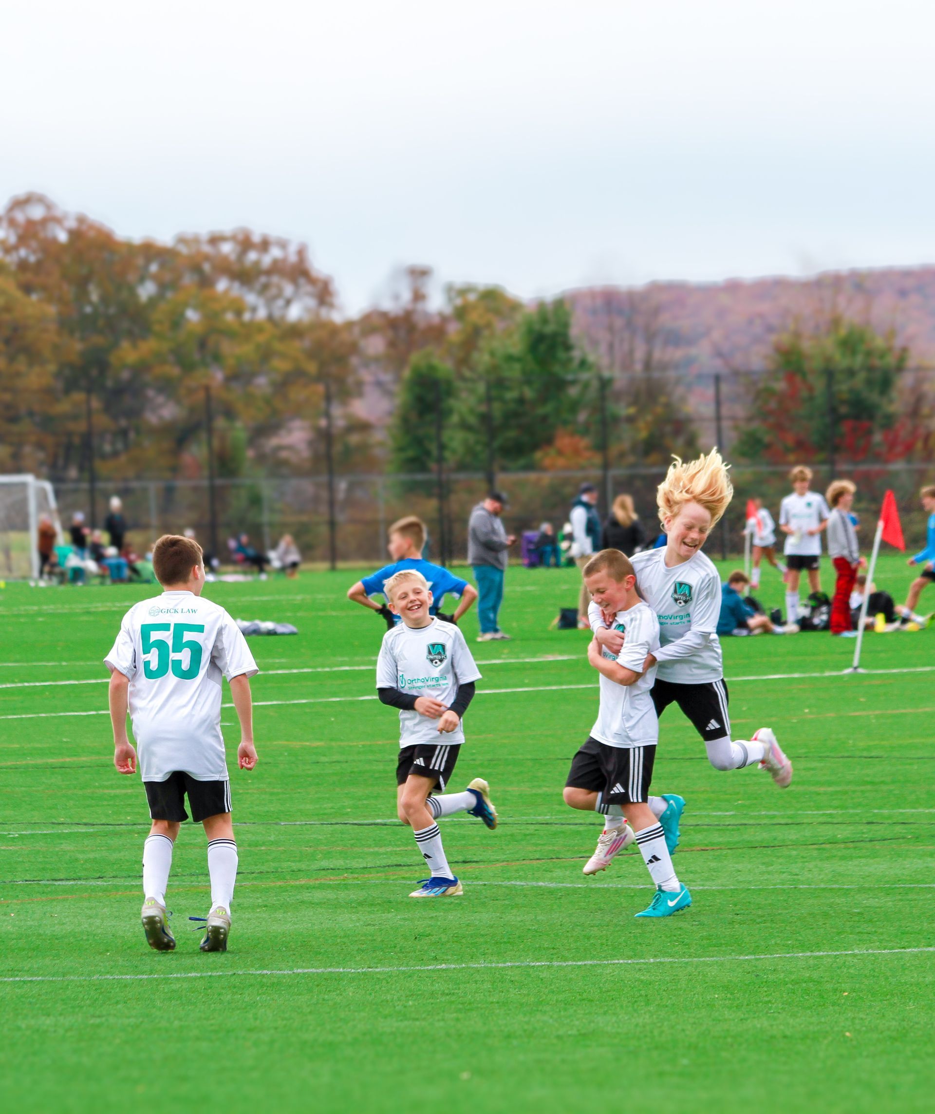 Soccer players celebrating on a green field. Two jump in excitement. Others run, wear white, teal, and black uniforms. Fall setting.
