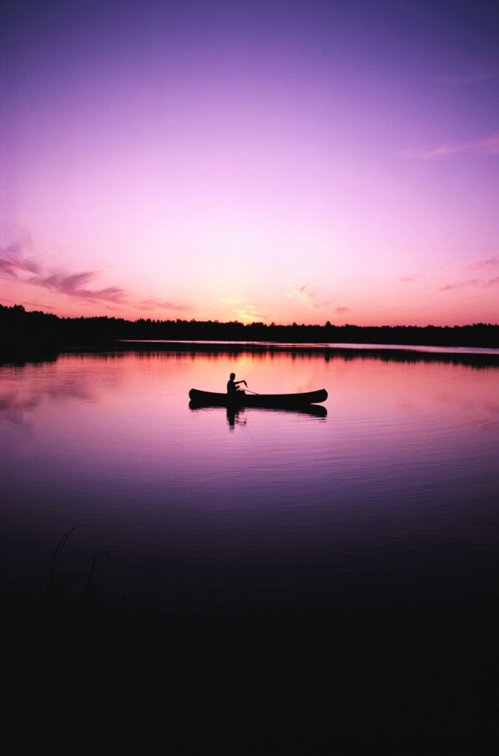 Silhouette of a person canoeing on calm water at sunset; purple and pink sky reflects on the lake.