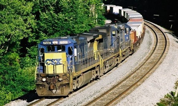 Blue and yellow CSX freight train rounding a bend on tracks through a green, wooded area.
