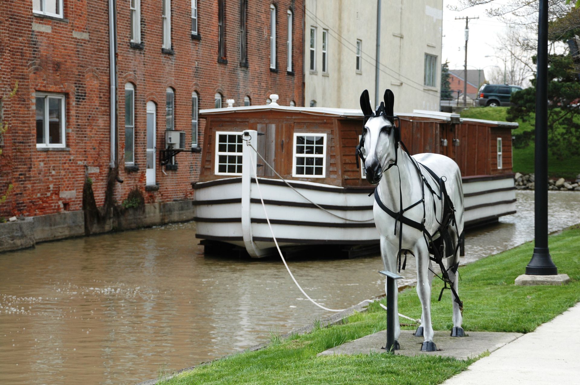 Horse-drawn canal boat on water; horse stands tethered on grassy bank next to brick building.