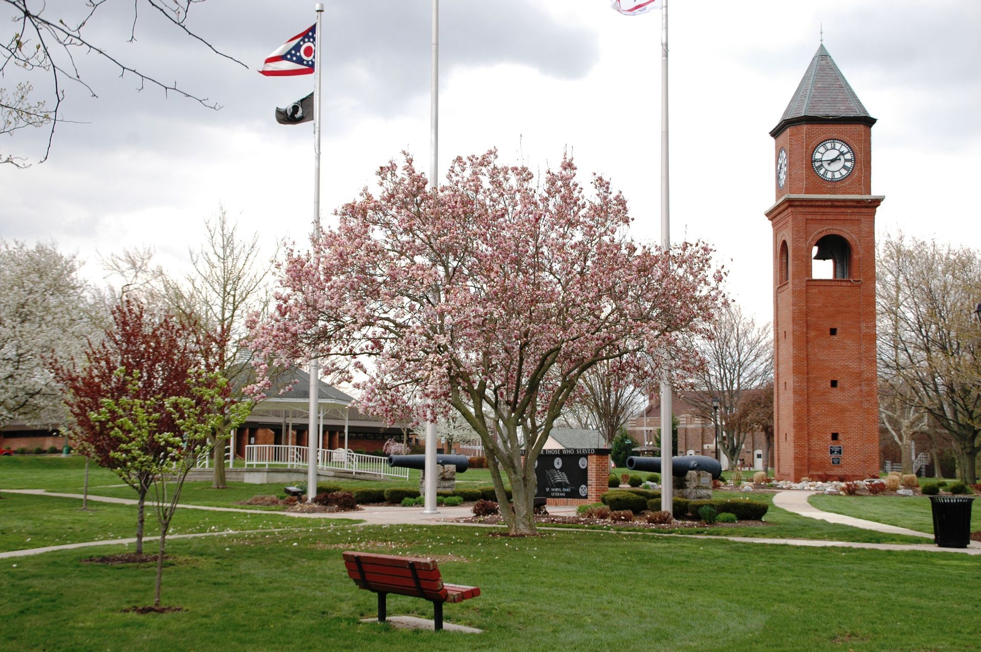 Park with blooming tree, clock tower, flags, gazebo, and bench.
