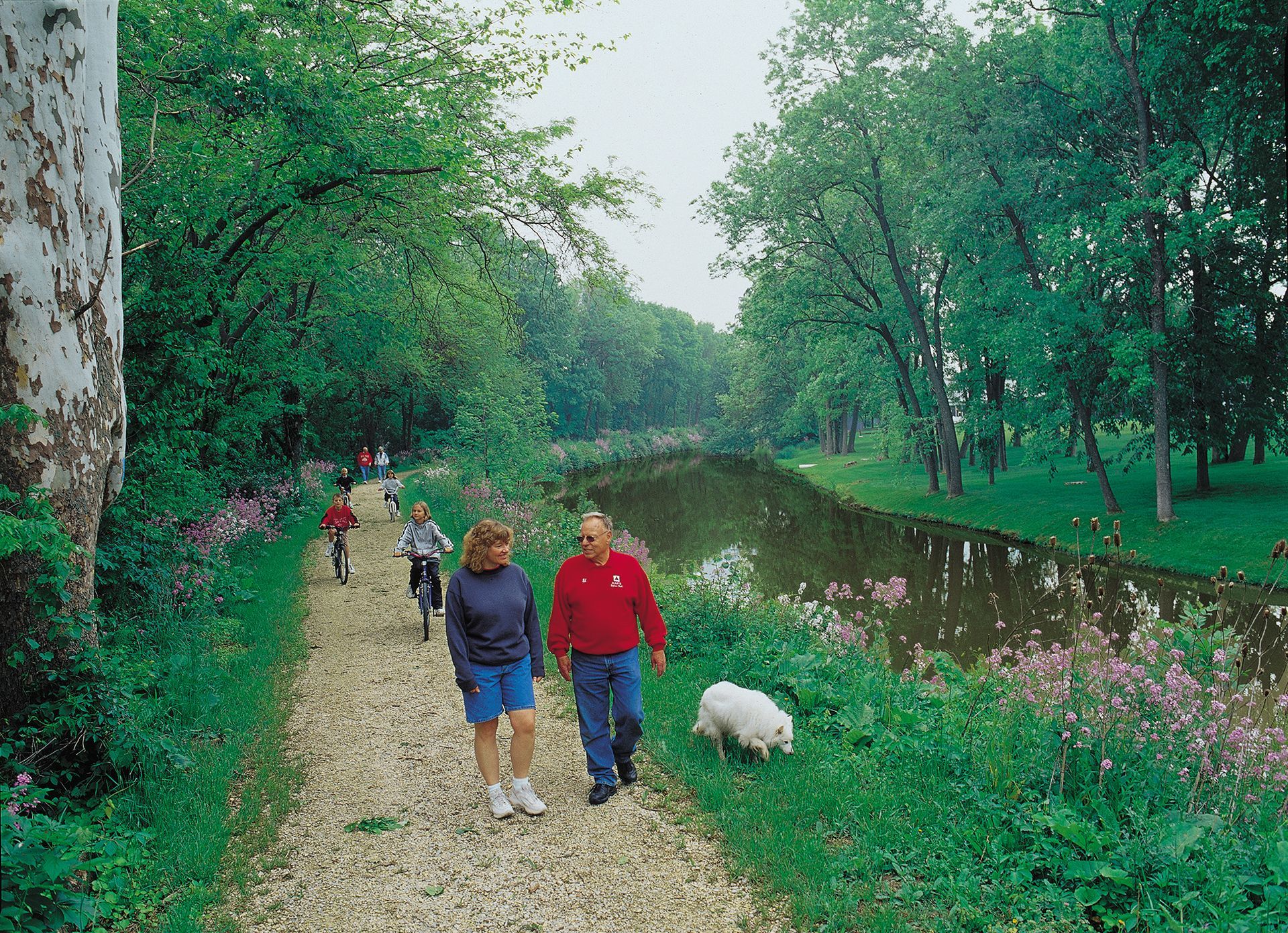 People walking on a gravel path along a canal, dog on leash, trees overhead.