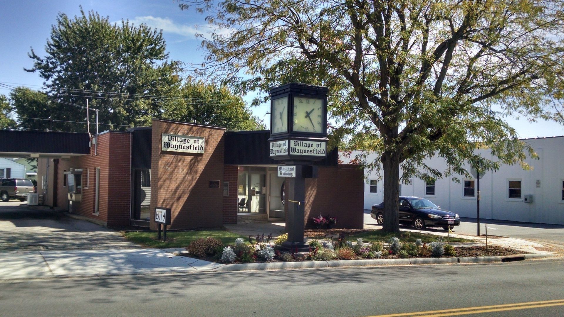 Clock tower in front of a brick building with a drive-through lane. A car and trees are in the background.